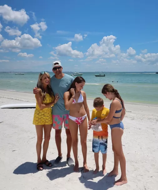 Family of five standing barefoot on a sunny beach in colorful swimwear, with boats on turquoise water and a clear sky behind them.